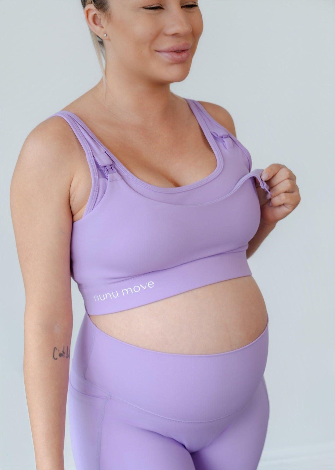 Close-up of a smiling pregnant mum wearing a Nunu Move activewear set in lilac. She demonstrates the easy-access, one-handed nursing clip on the breastfeeding sports bra for comfort and convenience.