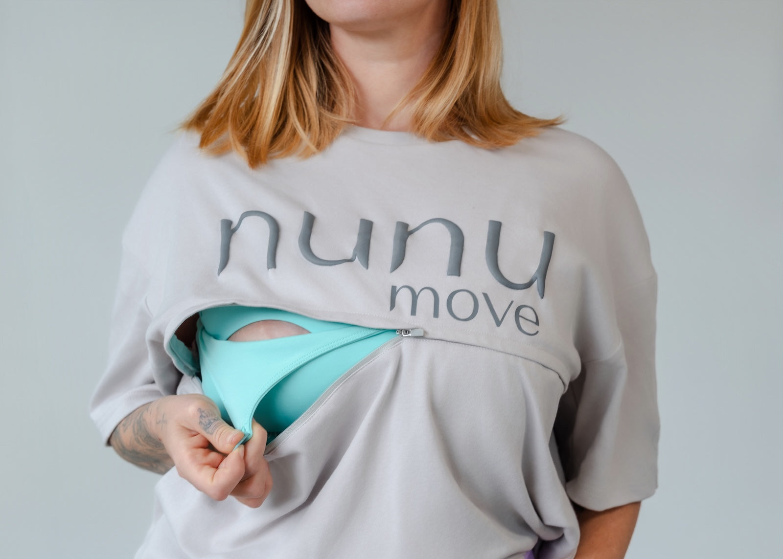 Close-up chest shot of a mum in the grey Nunu Move Oversized Nursing Tee, pulling open the zip access to reveal the teal bra underneath. The image highlights the logo and practical, quick nursing access of both the shirt and the nursing sports bra underneath.
