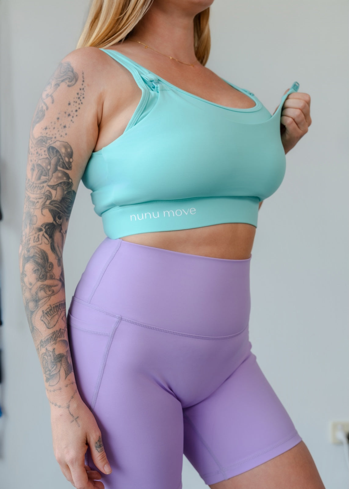 Close-up shot of a mum wearing Nunu Move breastfeeding activewear: a teal nursing sports bra and lilac active shorts. She demonstrates pulling the out layer to show the easy-access design.