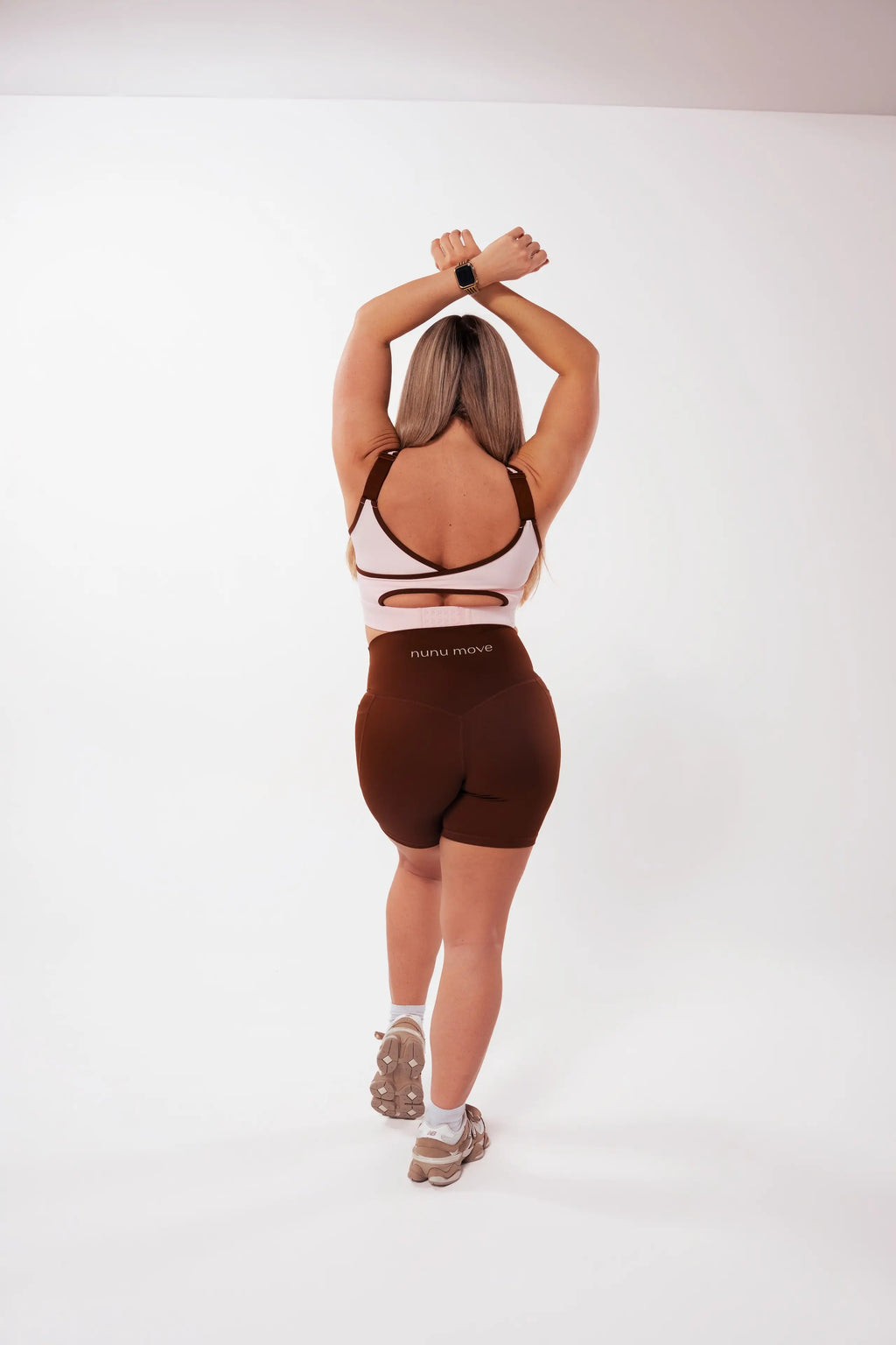 Back view of a woman wearing the Nunu Move Cherry Blossom Asta nursing bra with a strappy cut-out back, paired with Espresso brown V-Bum shorts, on a white studio background.