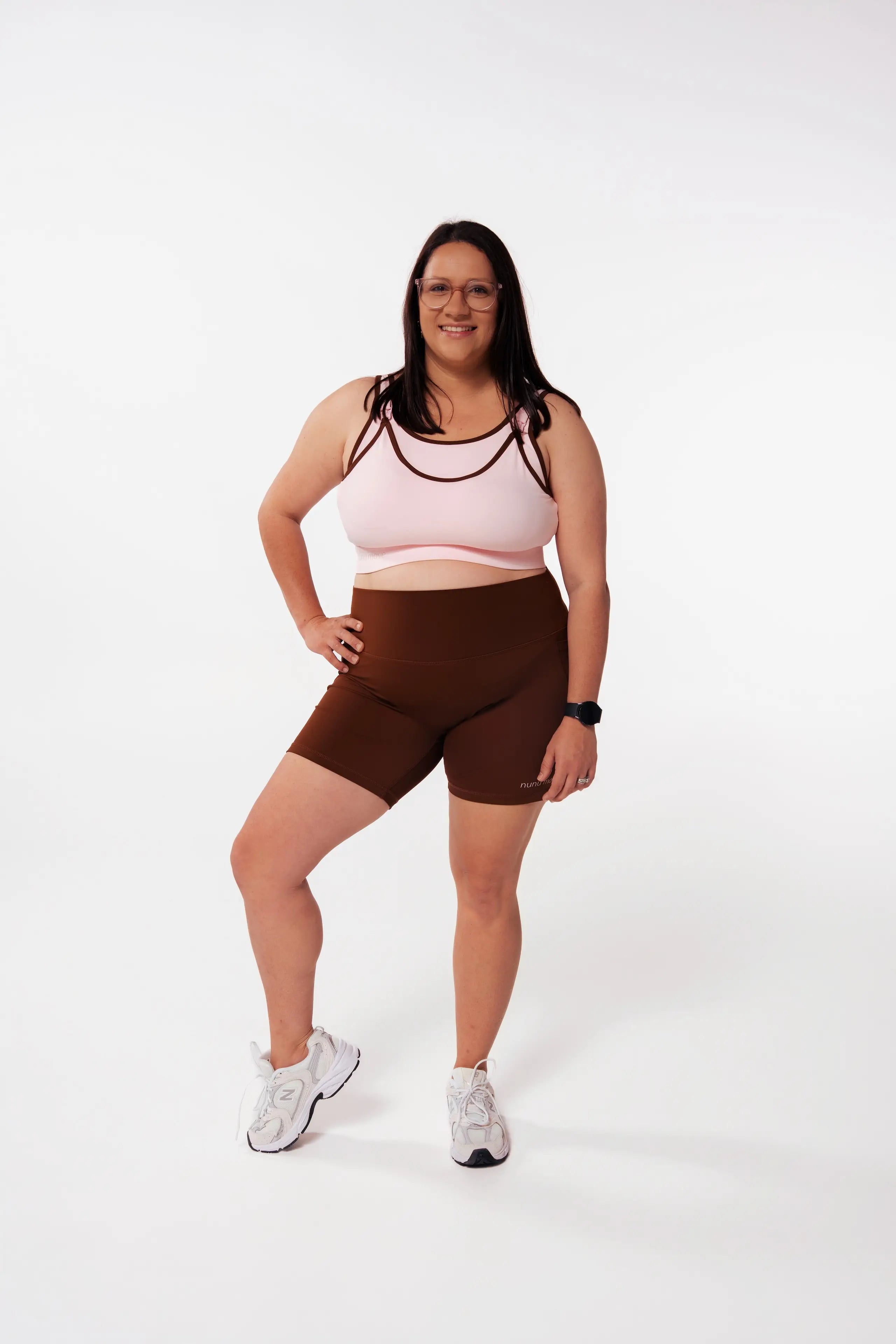 Woman wearing the Nunu Move Cherry Blossom Asta nursing bra with strappy detail and Espresso brown high-waisted bike shorts, front view on a white studio background.
