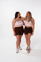 Two mums of different body types laughing together in Nunu Move Cherry Blossom Asta nursing bras and Espresso brown bike shorts, on a white studio background.