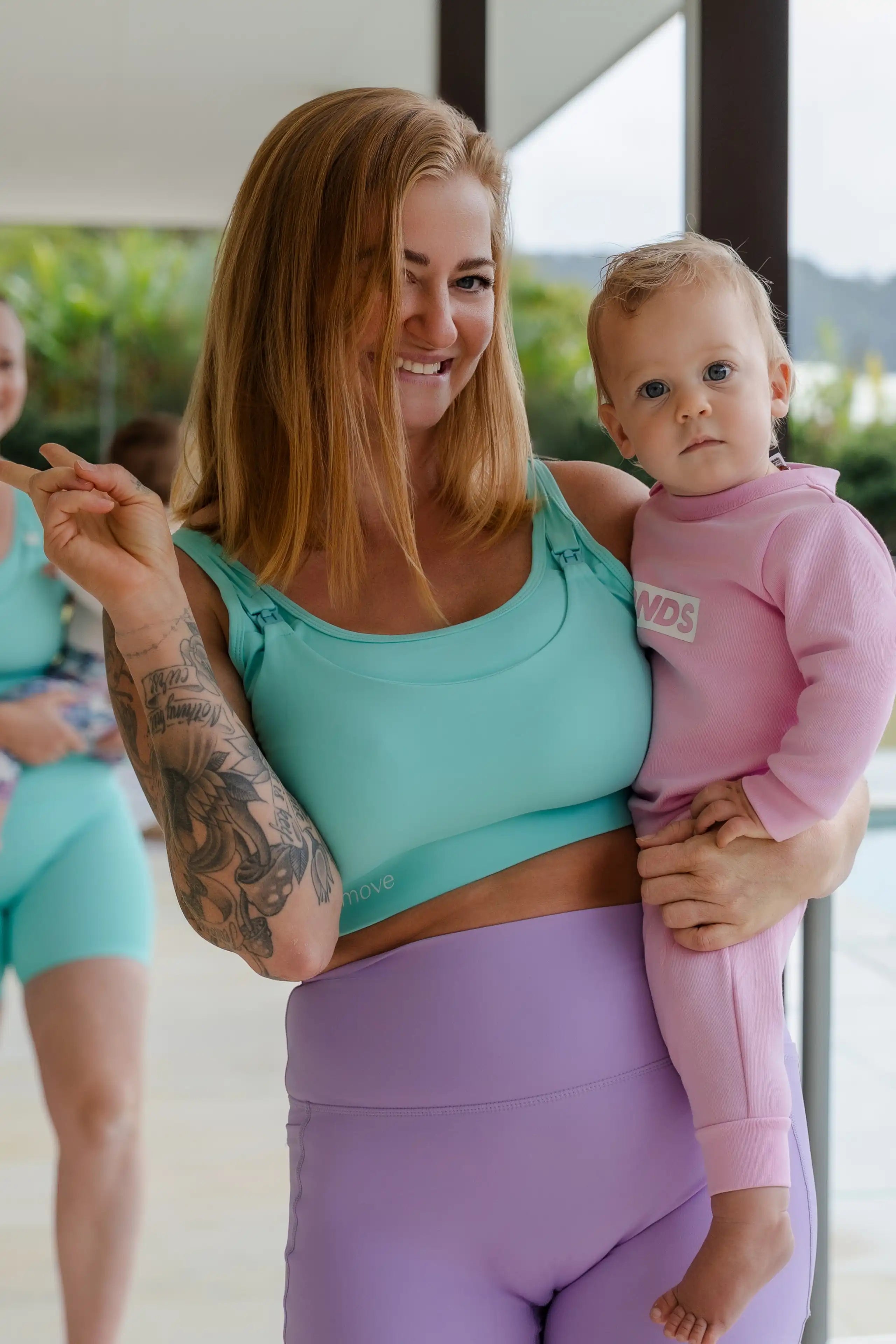 Nunu Move founder Shae smiling and holding her baby, wearing a mint nursing sports bra and lilac leggings on a Gold Coast patio.