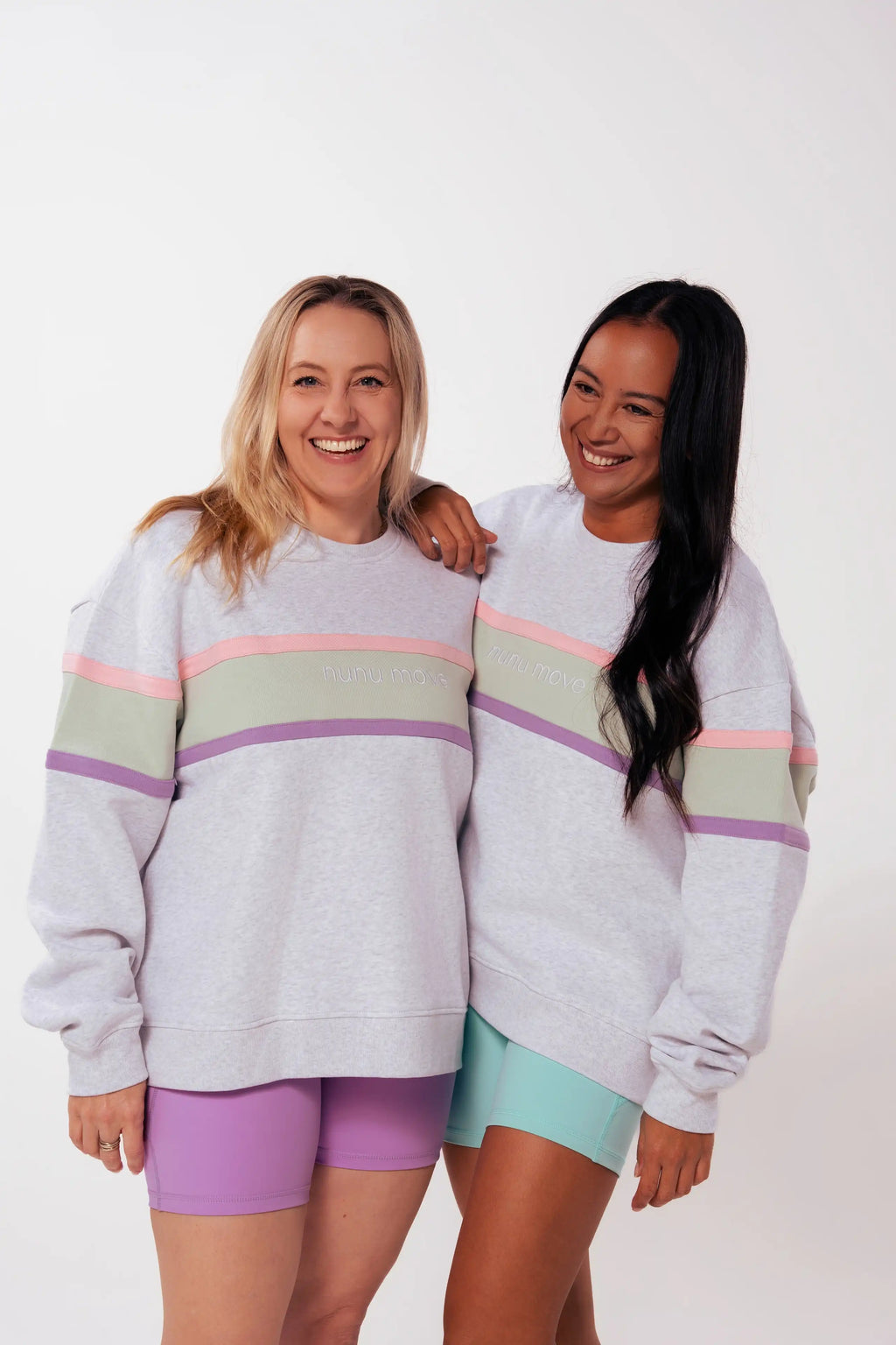 Two mums smiling in Nunu Move grey marle nursing jumpers on a white studio background, one in lavender bike shorts, one in mint bike shorts, showing the pink, sage and purple colourblock stripe and embroidered logo.
