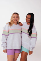 Two mums smiling in Nunu Move grey marle nursing jumpers on a white studio background, one in lavender bike shorts, one in mint bike shorts, showing the pink, sage and purple colourblock stripe and embroidered logo.