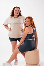 Two mums of different body types in Nunu Move activewear. One in the Oat oversized nursing tee with charcoal bike shorts, the other in a charcoal nursing bra and bike shorts set. On a white studio background.