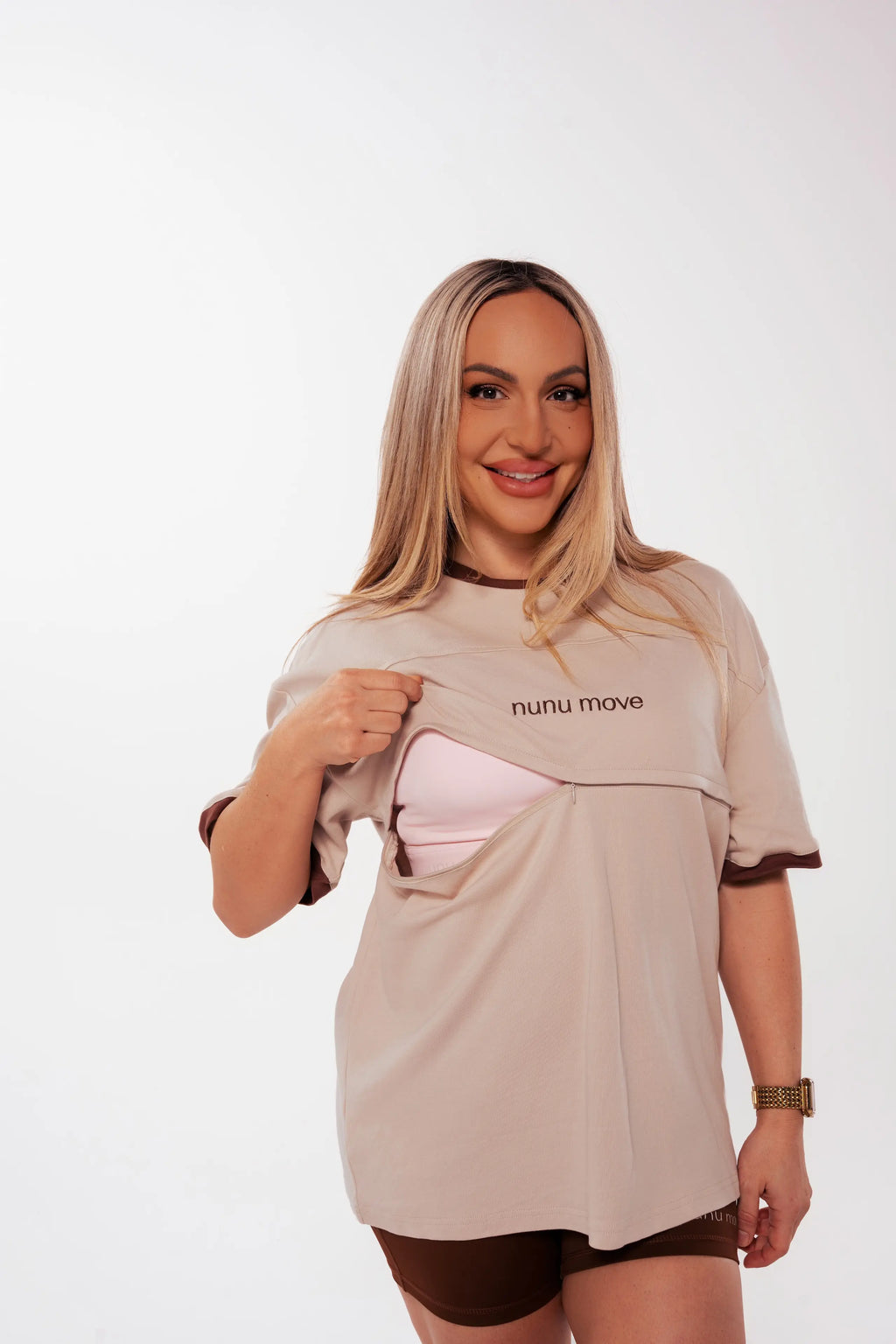 Smiling woman wearing the Nunu Move Oat oversized nursing tee, lifting the front panel to reveal the hidden zip feeding access over a pink nursing bra, on a white studio background.