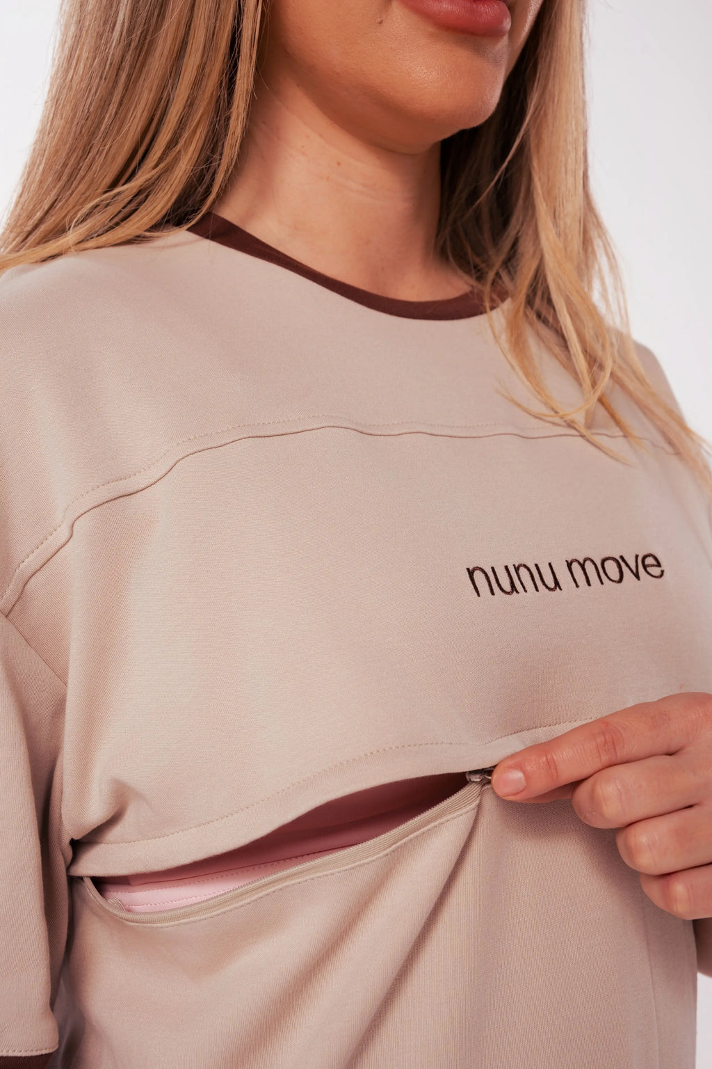 Close-up of the Nunu Move Oat nursing tee's horizontal zip feeding access opening below the embroidered nunu move logo, revealing a pink nursing bra.